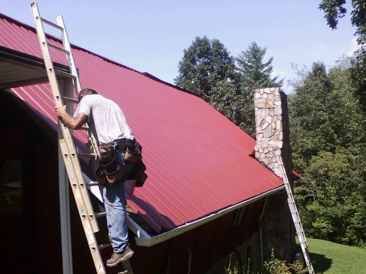 Roofer on a ladder beside a red metal roof and stone chimney — Soffit & Fascia Repair by Roofing Brothers in Fairbanks